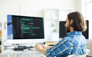 Person mit langen Haaren sitzt an einem Schreibtisch und tippt auf einer Tastatur vor einem großen Monitor, auf dem Code angezeigt wird.