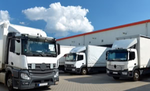 Mehrere weiße Lastwagen stehen auf einem gepflasterten Hof vor einem großen Gebäude mit roten Akzenten, bei blauem Himmel mit einigen Wolken.