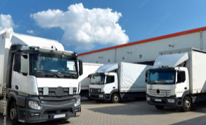 Mehrere weiße Lastwagen stehen auf einem gepflasterten Parkplatz vor einem großen Gebäude mit roten Dächern und Toren, unter einem blauen Himmel mit Wolken.
